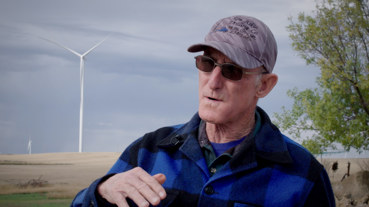 Man in baseball cap and flannel in front of field of wind turbines.