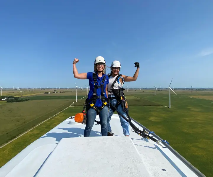 Two women flexing on top of a wind turbine wearing safety gear